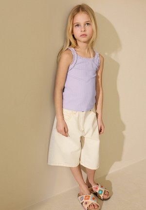 Jauna mergina su ilgais šviesiais plaukais vilki levandų spalvos be rankovių palaidinę, kreminius plačias kelnes šortus ir spalvingus nėrinius sandalų, stovinčią prie smėlio spalvos sienos.