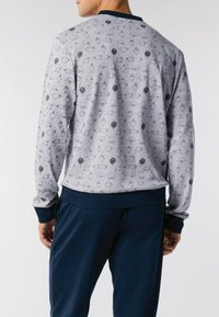 Uomo che indossa un felpa grigia chiara con polsini e vita blu scuro, decorata con piccole fantasie di uccelli cartone animato neri e grigi, abbinata a pantaloni blu scuro.