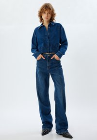 Camicia in denim tagliata con motivo floreale ricamato, tonalità blu chiaro, abbinata a jeans blu a gamba larga e scarpe nere a punta.
