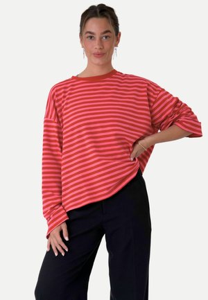 Mujer con una camisa de manga larga a rayas rojas y rosas y pantalones negros, de pie con una mano en la cadera sobre un fondo liso.
