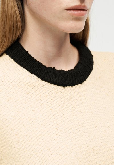 Gros plan sur une personne portant un pull en maille crème avec un col côtelé noir épais, montrant une partie du cou et du bas du visage.