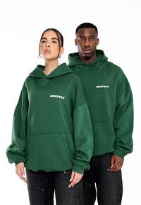 Sudaderas verdes de gran tamaño con un bolsillo frontal y el logo "MULTIPLY" en blanco. Presenta mangas relajadas y una textura acogedora. Llevada con jeans negros.