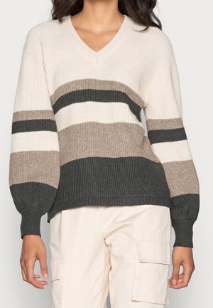 Femme portant un pull rayé à col en V crème, beige et gris foncé, associé à un pantalon cargo beige clair.