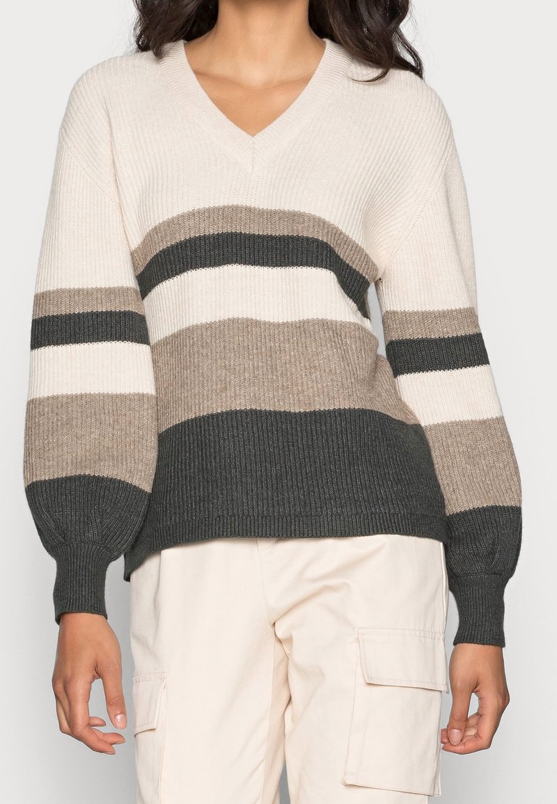 Femme portant un pull rayé à col en V crème, beige et gris foncé, associé à un pantalon cargo beige clair.
