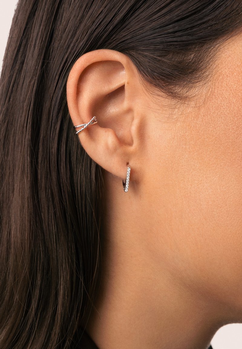Pendientes de aro plateados con piedras preciosas transparentes. Un pendiente es un aro delgado; el otro presenta un diseño retorcido, ambos montados en la oreja de un modelo.