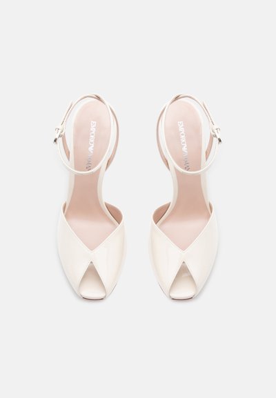 Λευκά λουστρίνι πέδιλα με ανοιχτά δάχτυλα, λουριά στον αστράγαλο και χαμηλά τακούνια, φαίνονται από πάνω σε απλό φόντο.