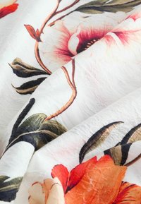 Tissu floral avec de grandes fleurs rouges et roses sur un fond blanc, ornées de feuilles vertes. La texture semble légèrement froissée.