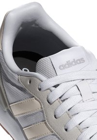 Weiße Sneaker aus Mesh- und Ledermaterialien, mit grauen Akzenten, strukturiertem Obermaterial, flachen Schnürsenkeln und einem gepolsterten Kragen mit Logo auf der Zunge.