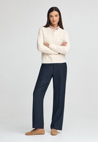 Cremfarbener Kabelstrickpullover mit gerippten Bündchen, marineblaue weit geschnittene Hosen und braune Wildlederloafers, ausgestattet mit einem schlichten runden Ausschnitt.