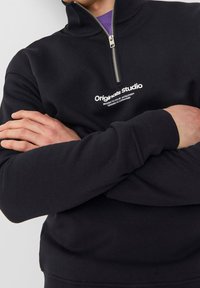 Černá sweatshirt na zip s vysokým límcem, na hrudi s bílým logem "Originals studio" a elastickými manžetami a lemem.