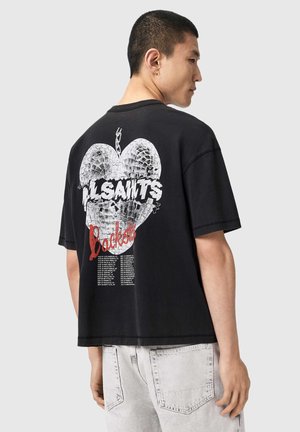 Jeune homme portant un t-shirt noir oversize avec un motif de cœur blanc éclaté et le texte « AllSaints Rockest » dans le dos, assorti à un jean denim clair.