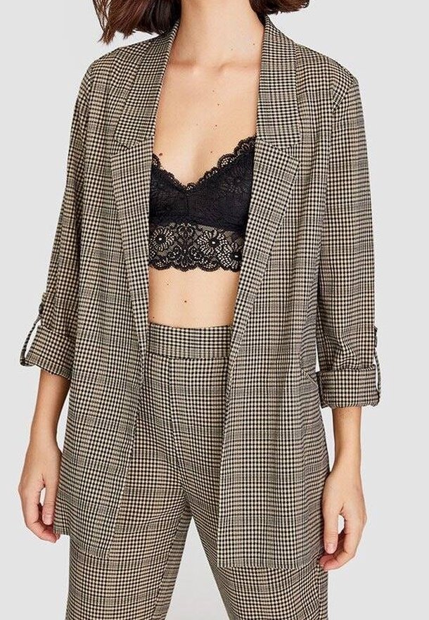 La femme porte un blazer à carreaux et un pantalon assorti avec les manches retroussées et un bralette en dentelle noire en dessous.