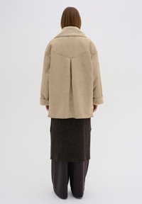 Béžová umělá kožichová bunda s širokým límcem, detailem větrání vzadu a texturovanými manžetami, nošená přes tmavohnědou vrstvenou sukni a široké kalhoty.