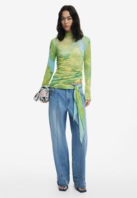 Top lungo attillato con maniche in verde e blu tie-dye, abbinato a jeans in denim larghi con un fiocco di stoffa coordinato. Tessuto texturizzato, design casual.