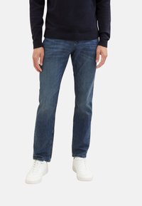 Jeans in denim blu scuro con taglio dritto, caratterizzati da un leggero sbiadimento. Indossati con sneaker bianche e un maglione di lana navy.