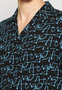 Chemise noire à manches courtes avec un motif répétitif en bleu imitant l'écriture manuscrite. Fermeture à boutons sur le devant avec col et poche poitrine.