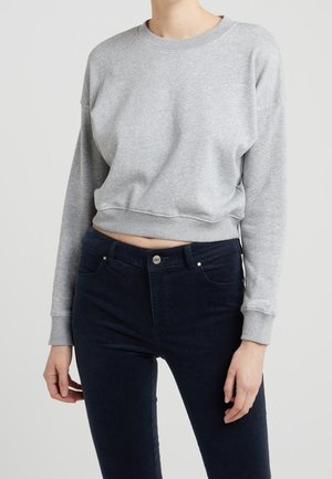 Person iført en lysegrå cropped sweatshirt og mørkeblå højtaljede jeans mod en ensartet lys baggrund.