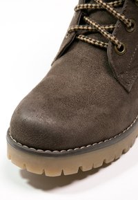 Bota de ante marrón hasta el tobillo con punta redonda, acabado texturizado, suela de goma y cordones a juego en contraste. Los detalles incluyen ojales de metal y costuras.