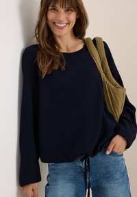 Blouse en maille côtelée navy à col rond et manches longues, dotée d'un ourlet cintré et d'un sac à épaule vert olive. Associée à un jean bleu clair.