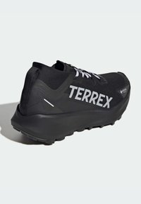 Crne trkaće tenisice s teksturiranim gornjim dijelom, bijelim vezicama i "TERREX" logom. Imaju robustan položaj za petu i izdržljiv potplat za prianjanje.