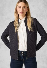 Veste matelassée noire avec des motifs géométriques, fermeture zippée complète et col montant. Portée sur une chemise blanche et un jean bleu. Texture lisse.