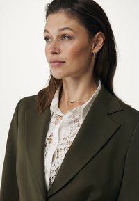 Blazer verde oliva sopra una blusa di pizzo bianco, con colletto classico, chiusura a bottoni e sottili dettagli in gioielleria dorata.