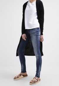 Svart lång kofta, vit texturerad V-ringad topp, blå skinny jeans och bruna sandaler med metallic detaljer och vävd sula.