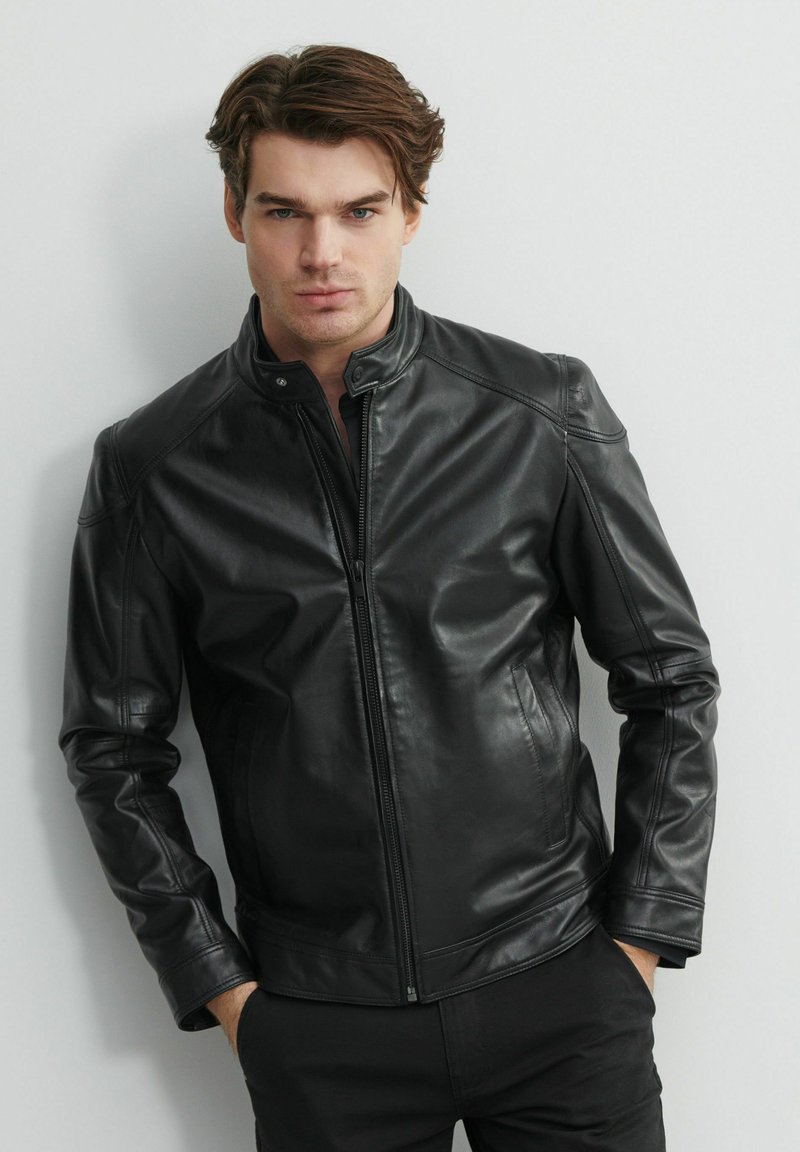Czarna skórzana kurtka z zapięciem na zamek, wysokim kołnierzem i dopasowanym krojem. Posiada gładką teksturę oraz przeszycia na ramionach.