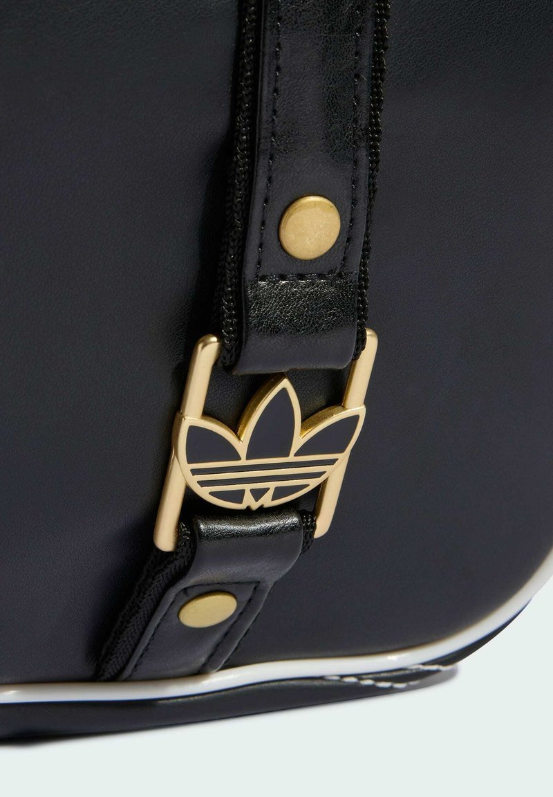 Sangle de sac en cuir noir avec boucle logo Adidas trefoil dorée et rivets dorés sur une surface lisse en cuir noir.