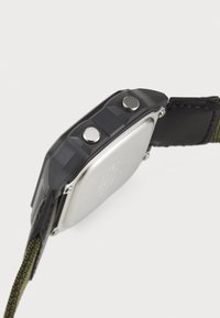 Montre noire avec un cadran rectangulaire en argent, deux boutons ronds sur le côté et un bracelet en tissu vert olive. Boîtier texturé pour une meilleure prise.