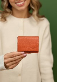 Brun læderpung med en tekstureret overflade, rektangulær form og et lille logo præget på forsiden, holdt i en hånd.