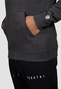 Dunkelgrauer Pullover mit Fronttaschen und gerippten Bündchen, mit einem kleinen weißen Logo am Ärmel. Kombiniert mit schwarzen Sweatpants.