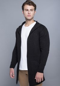 Sort hætte cardigan med en tekstureret strik, åben fortil, og to sidelommer. Sammen med en hvid t-shirt og khaki bukser.