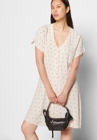 Vestido de manga curta, com decote em V, em bege claro com bolinhas pretas, apresentando uma frente com botões e um corte descontraído. Modelo segura uma pequena mala preta.