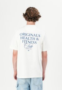 Personne avec des cheveux bouclés portant un t-shirt blanc avec le texte "Originals Health & Fitness Club Since 1972" dans le dos et un short en denim bleu clair.