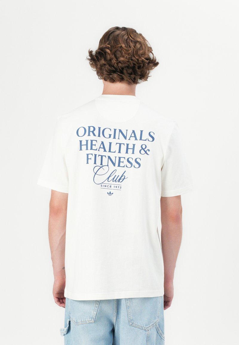 Personne avec des cheveux bouclés portant un t-shirt blanc avec le texte "Originals Health & Fitness Club Since 1972" dans le dos et un short en denim bleu clair.