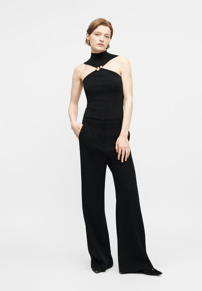 Γυναίκα που φοράει μαύρο ασύμμετρο τοπ χωρίς μανίκια με χρυσά κουμπιά και μαύρο παντελόνι φαρδύ, στέκεται με το ένα χέρι στην τσέπη.