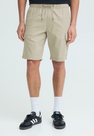 Mann iført beige cargoshorts med snor i livet, hvite crew-sokker og svarte Adidas-sneakers med hvite striper.