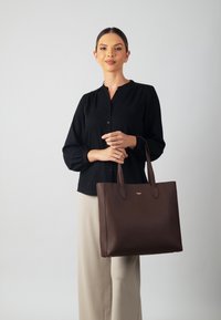 Brown leather tote bag with textured surface and dual handles. Features a minimalist design and a small logo detail on the front.