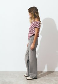 Femme se tenant de côté, portant un haut à rayures avec épaules dénudées, un pantalon gris ample taille haute avec des clous sur le côté, et des chaussures blanches.
