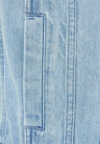 Světle modrá džínová látka s viditelnou texturou a prošitím. Na přední straně má náprsní kapsu, která zvyšuje její užitečnost a design.