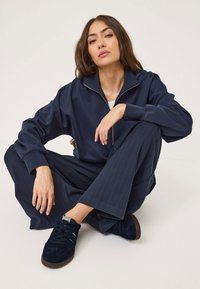 Mornarska zip-up dukserica z rebrastimi manšetami, v kombinaciji z širokimi hlačami s črtami in temnimi semenskimi supergami, kar ustvarja usklajen videz.