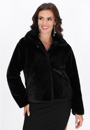Frau, die lächelt, trägt eine schwarze Kunstpelzjacke und lange Kristall-Ohrringe, steht vor einem weißen Hintergrund.