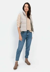 Hellbeige gefütterte Weste mit hohem Kragen, strukturierter beiger Strickpullover, blaue Jeans und braune Stiefeletten, stehend vor einem neutralen Hintergrund.