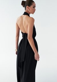 Črna obleka s halter vrvico in odprtim hrbtom, z detajlom zavezovanja v pasu. Gladek material s pretočno silhueto. Doposrena z izrazitimi uhani.