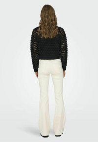 Schwarzer, gestrickter, verkürzter Pullover mit offenem Muster, kombiniert mit weißen, ausgestellten Hosen. Das Outfit hat einen enganliegenden Bund und lange Ärmel.