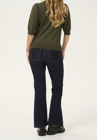 Kvinna med långt vågigt blont hår, klädd i en grön kortärmad topp, mörkblå bootcut-jeans och svarta klumpiga skor, som står vänd bort.