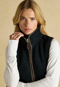 Schwarze Fleece-Weste mit hohem Kragen, ausgestattet mit einem braunen Reißverschluss und Akzenten. Getragen über einem weißen Langarmshirt, das einen strukturierten Stoff zeigt.