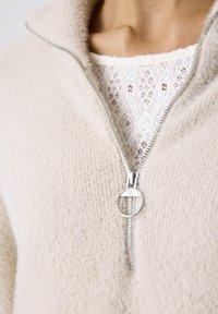 Close-up of beige fuzzy zip-up sweater with silver ring zipper pull, worn over a white lace-knit top at the neckline.