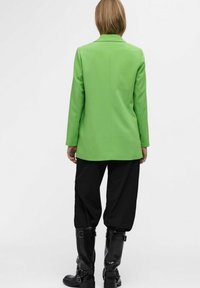 Groene op maat gemaakte blazer met lange mouwen, een kraag en een recht model, gecombineerd met zwarte, loszittende broeken en zwarte laarzen tot de knie.
